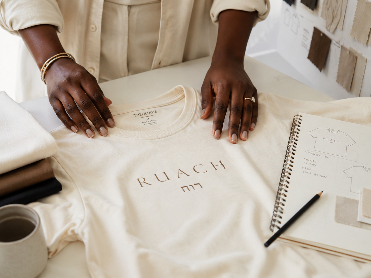 A cream-colored t-shirt with 'RUACH' and Hebrew lettering printed on the front, styled on a table with a person's hands resting on it, alongside a notebook, pencil, and folded garments.